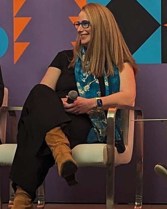 Beth Rudden seated on a panel stage at SXSW EDU, holding a microphone, in her Van Gogh almond-blossom scarf and tan suede boots.